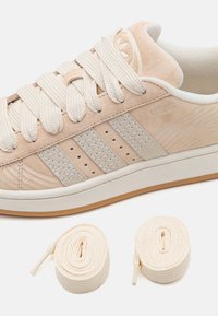 Beige suede sneaker with three textured side stripes, flat cream laces, and a rubber sole. Two additional laces in matching color included.