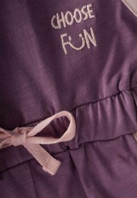 Mono de algodón morado con cintura elástica, que presenta un cordón rosa y texto bordado "ELIGE DIVERTIDO". Textura suave y diseño casual.