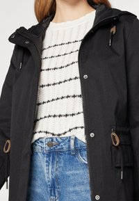 Svart lättviktsjacka med dragkedja och huva. Har bruna togglar som detaljer och en framficka. Bärs över en vit mönstrad tröja.