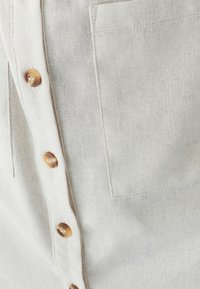 Jasno beżowa koszula z teksturowanej tkaniny, z rzędami okrągłych brązowych guzików oraz naszywaną kieszenią po lewej stronie.