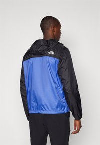 Giacca a vento blu e nera con cappuccio; presenta una texture liscia e colori a contrasto; logo sulla schiena.