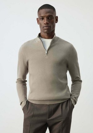Man draagt een beige trui met kwart rits over een wit overhemd en donkerbruine broek, staat met handen in de zakken tegen een effen achtergrond.