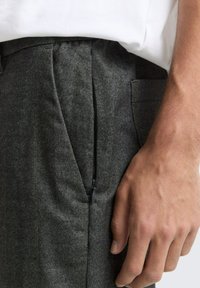 Hand ruht neben dunkelgrauen, plissierten Hosen mit einem dezenten seitlichen Reißverschluss und einem darin gesteckten weißen Hemd.