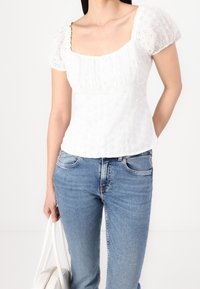 Blusa di cotone bianca con maniche a sbuffo, scollatura quadrata e motivo a occhielli floreali. Abbinata a jeans in denim blu e una borsa bianca.