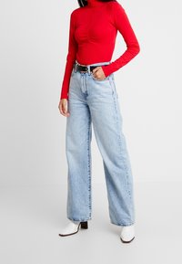 Frau in einem roten Rollkragenpullover, hellblauen, hochgeschnittenen, weiten Jeans mit einem schwarzen Gürtel und weißen Stiefeletten, die vor einem weißen Hintergrund steht.
