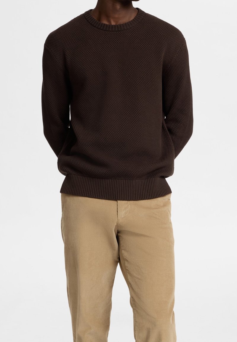 Suéter marrón texturizado con cuello redondo, puños y dobladillo acanalados, combinado con pantalones de pana caqui. La tela presenta un patrón de diamantes.