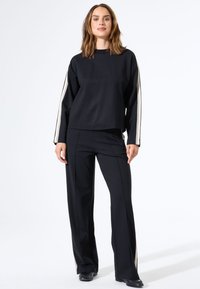 Sudadera negra corta con mangas largas que presenta rayas laterales en color crema, combinada con pantalones negros ajustados con detalle de raya lateral.