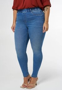 Blauwe denimleggings met een taps toelopende pasvorm, rekbare tailleband en subtiele vervaging. Gecombineerd met een losse rode blouse en naaktkleurige sandalen met banden.