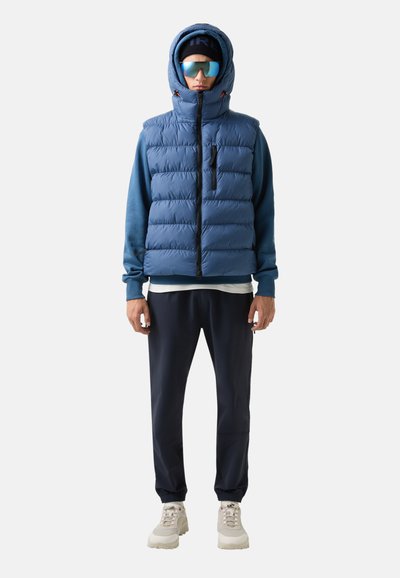 Blaues Steppwesten mit hohem Kragen und Kapuze, getragen über einem blauen Sweatshirt, kombiniert mit navyfarbenen Hosen und cremefarbenen Sneakers; inklusive Sonnenbrille.