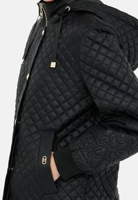 Chaqueta acolchada negra con capucha, detalles dorados, cierre de cremallera y puños texturizados. Relieve floral distintivo en la manga.