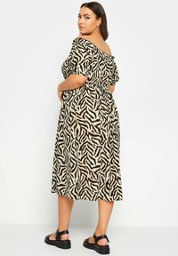 Abito midi off-the-shoulder beige con strisce zebrate nere, caratterizzato da una vita arricciata e maniche a volant, abbinato a sandali neri.