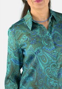Blusa verde con estampado paisley y acentos en azul y marrón, con cuello tipo camisa, mangas largas y cierre frontal de botones. Tejido suave y brillante.