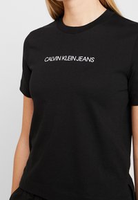 Czarny t-shirt z krótkim rękawem z białym napisem "CALVIN KLEIN JEANS" na piersi, noszony przez osobę o jasnej skórze i blond włosach.