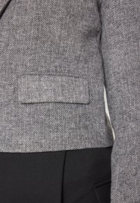 Šedá látka s herringbone vzorem na saku s viditelnou kapsou, doplněná o jednobarevný černý materiál, který zvýrazňuje přizpůsobený styl.