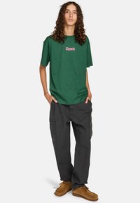 Groene katoenen T-shirt met korte mouwen, voorzien van een roze "Element" logo, gecombineerd met donkergrijze cargobroeken en bruine veterschoenen.