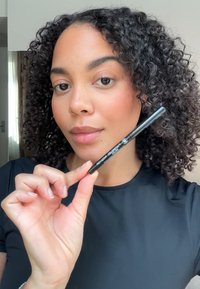 Zwarte eyelinerpotlood vastgehouden in de hand, cilindervorm, matte afwerking. Draagt een persoon met krullend haar, gekleed in een zwarte top, neutrale achtergrond.