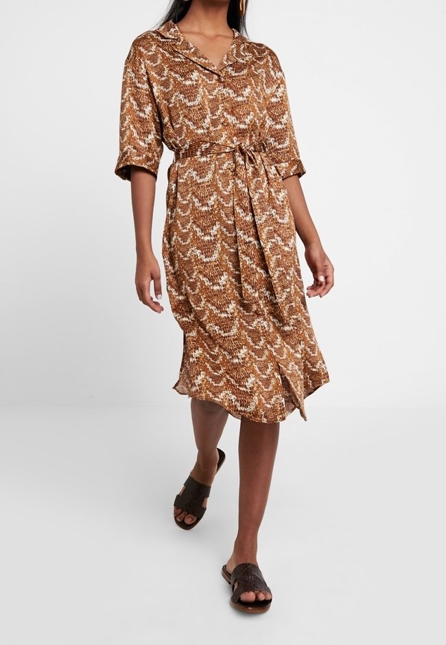 Femme portant une robe marron à motifs longueur genou avec une ceinture à la taille et des manches trois-quarts, marchant avec des sandales sombres à enfiler.