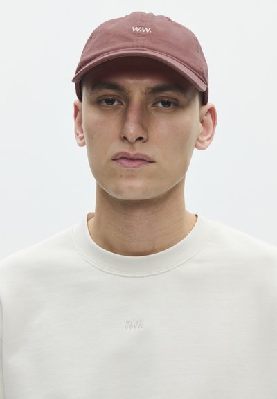Jonge man met een gedempt roze pet met het logo "W.W." en een gebroken witte sweatshirt met een subtiel geborduurd "W.W." op de borst.
