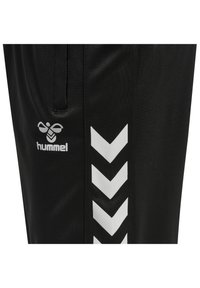 Svarta träningsbyxor med en slät yta, försedda med en vit Hummel-logotyp och ett vitt chevron-mönster längs den yttre sidan.