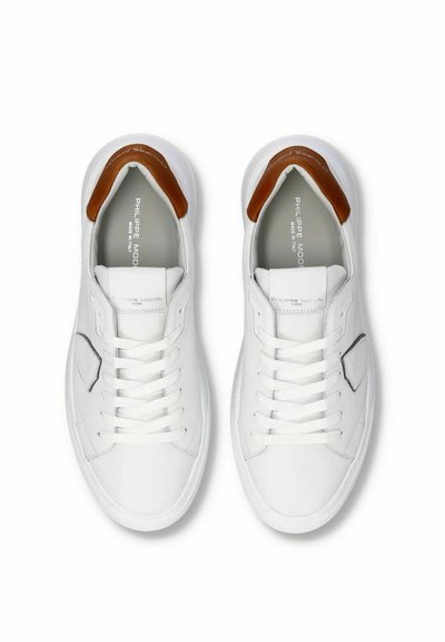 Hvide lædersneakers med en brun accent ved hælen, med snørebånd foran, syninger og et minimalistisk design.