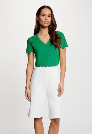 Vrouw in een groen V-hals top met kanten mouwen met oogjes en witte shorts tot op de knie, staand tegen een effen lichte achtergrond.