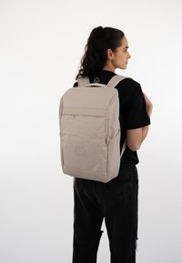 Light beige backpack with a smooth texture and a front zipper pocket, featuring adjustable straps and rectangular shape. Minimalist design.