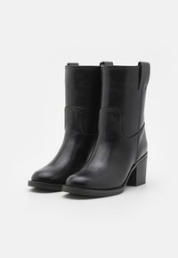 Par de botas de cuero negras hasta medio pantorrilla con puntas redondeadas, tacones apilados y lengüetas, exhibidas sobre un fondo blanco simple.