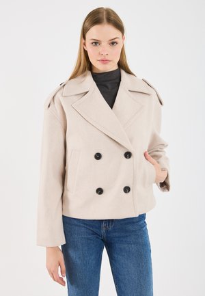 Ung kvinna med en beige dubbelknäppt kappa över en svart polo-tröja och blå jeans, som står framför en enfärgad vit bakgrund.