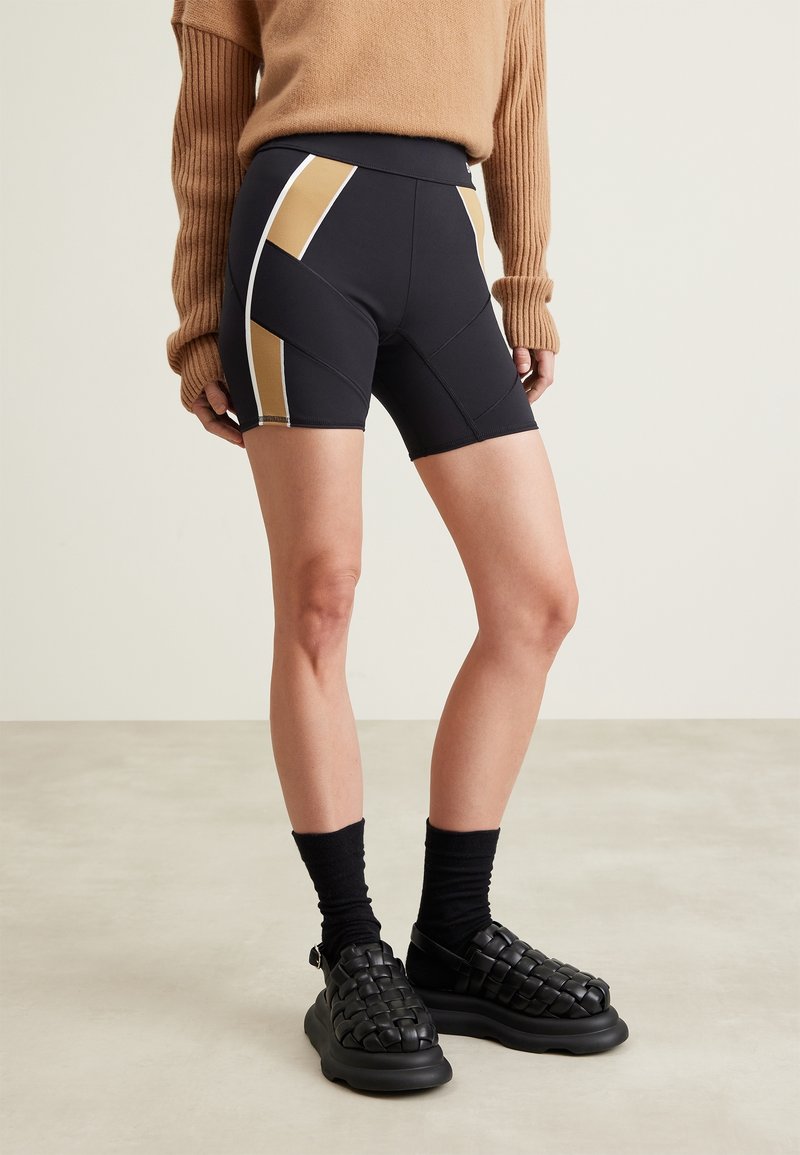 Personne portant un short de cyclisme ajusté noir et beige, des chaussettes noires, des sandales tissées noires et un pull marron à manches longues, debout sur un sol uni.