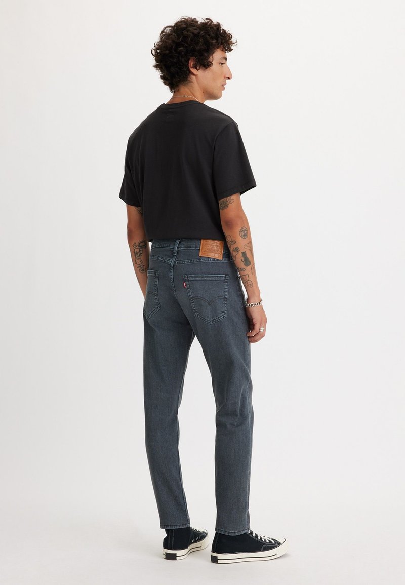 Dunkelblaue Jeans mit gerader Passform, Leder-Patch auf der Rückseite, kombiniert mit einem schwarzen T-Shirt und schwarzen Sneakers, von hinten gezeigt.