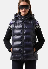Frau trägt übergroße schwarze Sonnenbrille, eine glänzende marineblaue gesteppte ärmellose Jacke und einen schwarzen Pullover mit gemusterten Schultern.