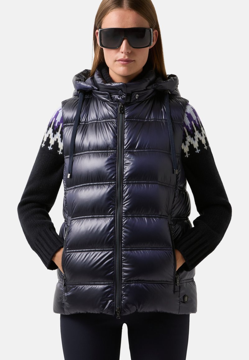 Frau trägt übergroße schwarze Sonnenbrille, eine glänzende marineblaue gesteppte ärmellose Jacke und einen schwarzen Pullover mit gemusterten Schultern.
