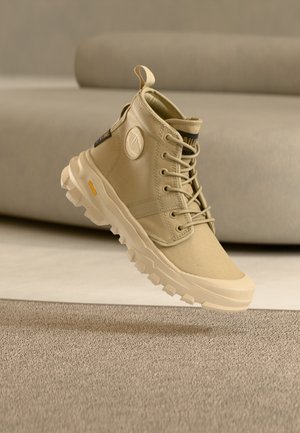 Smėlio spalvos aukštos viršutinės drobinės sportbačiai suapvalintu logotipo elementu, raištelių užsegimu ir tvirtu padu su matomais sukibimo raštais.