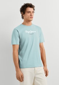 Άντρας που φοράει ανοιχτό μπλε μπλουζάκι Pepe Jeans London και λευκό παντελόνι, στέκεται μπροστά σε ένα απλό ανοιχτό γκρι φόντο.