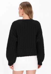 Čierny pletený sveter s textúrovaným vzorom, so spustenými ramennými švami a rebrovaným lemom. Spárovaný s bielou sukňou, ktorá zdôrazňuje skrátený strih.
