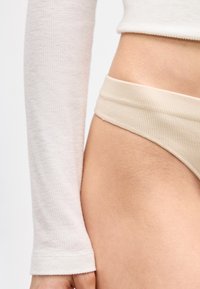 Κοντινό πλάνο στον κορμό και το χέρι ενός ατόμου που φοράει μακρυμάνικο ριμπ τοπ σε κρεμ απόχρωση και μπεζ ριμπ σλιπ ψηλού κοψίματος.