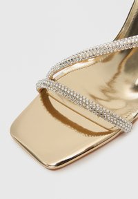 Sandale plată aurie cu două barete decorate cu pietre strălucitoare clare și vârf pătrat. Suprafață netedă cu finisaj reflectorizant.