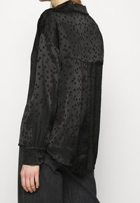 Svart, semi-genomskinlig blus med prickigt mönster, långa ärmar och krage. Har knapppynt på ärmsluten och en lös passform.
