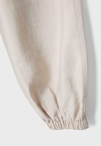 Jambe de pantalon en tissu blanc cassé avec poignet élastique et couture latérale visible sur un fond clair.
