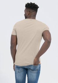 Beżowa dopasowana koszulka z krótkim rękawem, gładka w dotyku, okrągły dekolt i zaokrąglony dół. Nosić z niebieskimi dżinsami. Widok z tyłu.