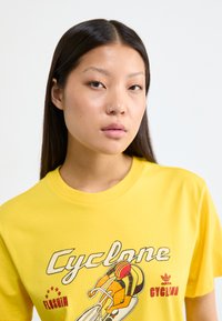 Yellow cotton t-shirt featuring a cycling graphic with a cyclist in orange and red. Text reads "Cyclone" and "adidas CYCLING." Short sleeves.