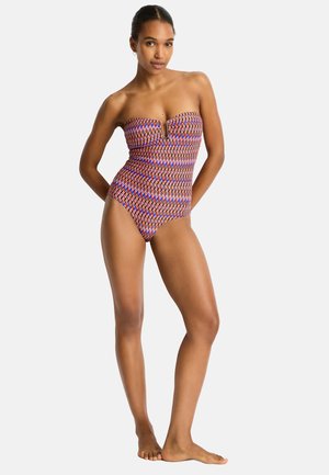Femme debout pieds nus portant un maillot de bain une pièce sans bretelles avec des formes géométriques en bleu, orange et blanc.