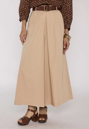 Donna che indossa pantaloni beige a gamba larga con cintura marrone, camicetta marrone a pois, braccialetti dorati e sandali marroni con tacco.