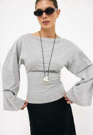 Mujer con gafas de sol negras, una camiseta gris holgada de manga larga, una falda negra y un collar largo con un colgante plateado.