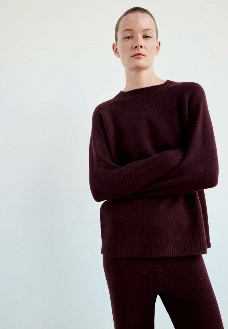 Femme aux cheveux courts portant un pull à manches longues bordeaux foncé et un pantalon assorti, debout les bras croisés devant un fond uni.