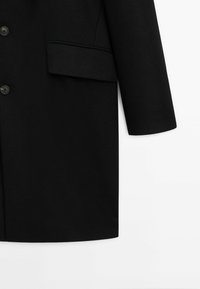 Manteau en laine noire avec poche à rabat, trois boutons noirs et manches longues droites sur fond blanc.
