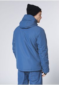 Blaue isolierte Jacke mit Kapuze, strukturiertem Stoff, elastischen Bündchen und Rückenabluftung. Wird mit einer schwarzen Mütze getragen und zeigt ein schlankes Design.