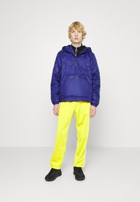 Giacca imbottita viola con cappuccio e mezza zip, abbinata a pantaloni gialli brillanti e sneakers nere. Texture liscia e colori a contrasto.