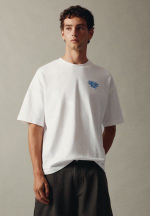 Jeune homme aux cheveux bouclés portant un t-shirt blanc ample avec un petit logo bleu "Surfider Beach" et un pantalon foncé sur un fond uni.