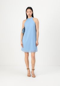 Light blue halter dress with a sheer overlay and subtle polka dot pattern; model holding a small blue clutch; tan strappy sandals.